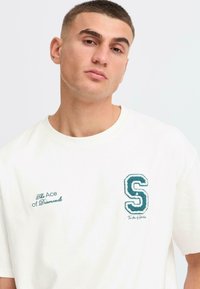 White cotton t-shirt featuring green embroidered accents, including the letter "S" and text "The Ace of Diamonds" on the front.