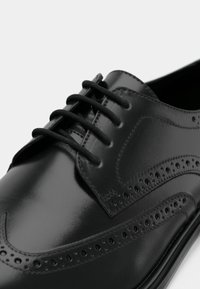 Shoe brogue preto em couro com perfurações decorativas e atacadores pretos, apresentado em close-up sobre um fundo branco.