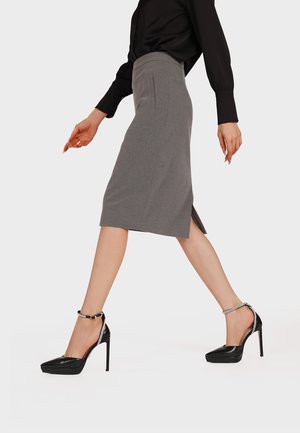 Donna che cammina indossando una gonna a tubino grigia al ginocchio, una camicetta nera e scarpe nere con tacco alto e cinturino alla caviglia, su uno sfondo neutro.