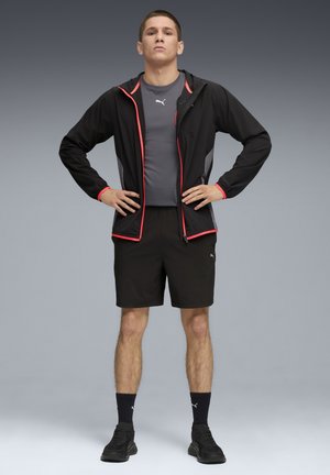 Sort letvægtsjakke med røde detaljer, grå atletisk skjorte, sorte shorts og sorte sneakers. Modellen står med hænderne på hoften.