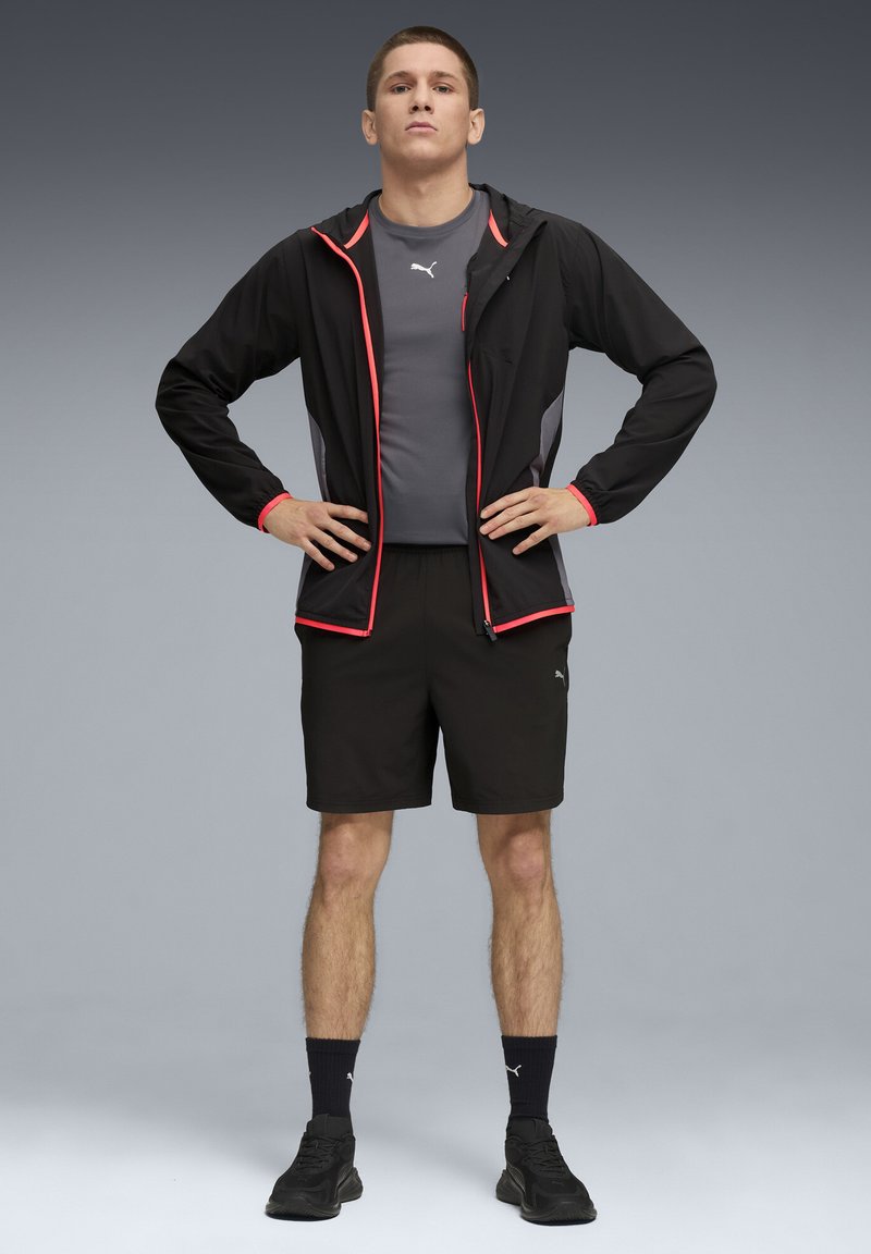 Chaqueta ligera negra con acentos rojos, camiseta atlética gris, pantalones cortos negros y zapatillas negras. El modelo está de pie con las manos en las caderas.