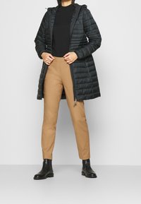 Doudoune noire avec capuche, dotée d'une fermeture éclair, de coutures horizontales et de poches latérales, associée à un pantalon slim beige et des bottes noires.