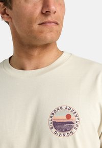 Camiseta color beige claro hecha de algodón, con un gráfico circular que presenta un diseño de atardecer en múltiples colores y el texto "BILLABONG ADVENTURE DIVISION".