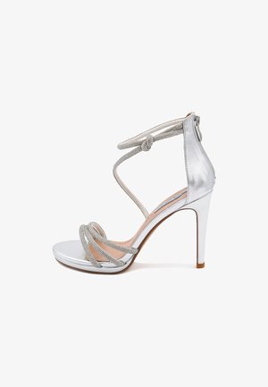 Sandal con tacco alto argento, caratterizzata da una tomaia metallica liscia, diverse cinghie decorative e un dettaglio annodato alla caviglia con chiusura a zip.