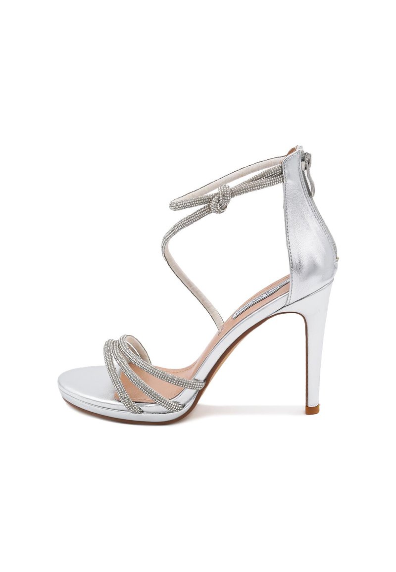 Sandal con tacco alto argento, caratterizzata da una tomaia metallica liscia, diverse cinghie decorative e un dettaglio annodato alla caviglia con chiusura a zip.