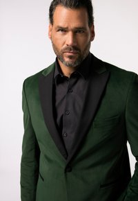 Blazer verde in velluto con revers in satin nero, chiusura con un bottone e fazzoletto da taschino; indossato su una camicia nera con bottoni.