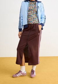 Osoba oblečená v světle modré bundě Adidas, květinovém topu, hnědé sukni Adidas s růžovými pruhy a předním zipem, bílých ponožk ách a fialových teniskách Adidas.