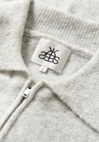 Svetlo siva zip-up jopica z ribanim ovratnikom. Narejena iz mehkega materiala, z oznako z logotipom in oznako velikosti "M."