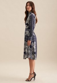 Abito blu navy con motivi paisley bianchi, maniche lunghe, collo alto e orlo svasato, abbinato a sandali neri con tacco alto.