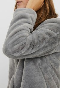 Šedá umělá kožešinová bunda s žebrovanou strukturou. Detailní záběr na rukáv, kter�ý ukazuje měkkou látku a mírně nadýchaný siluetu. Žádné viditelné kovové prvky.