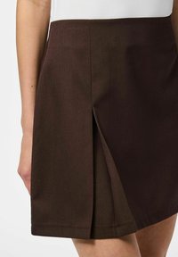 Jupe tailleur marron avec un subtil détail de pli à l'avant, fabriquée en tissu lisse, dotée d'un ourlet net et sans fermetures visibles.