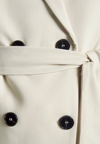 Krämfärgad blazer med textured tyg, utrustad med två svarta knappar och en knytdetalj i midjan.
