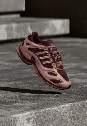 Zapatilla de correr granate y rosa con parte superior de malla y sintética flotando sobre escalones de hormigón gris, proyectando una sombra debajo.