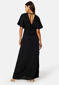 Abito maxi nero con maniche corte ampie, schiena scoperta con chiusura a fiocco e texture liscia. Lungo e fluido, con tasche laterali.