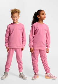 Zwei Kinder tragen passende pinke Sweatshirt- und Jogginganzug-Sets aus glattem Stoff. Die Outfits haben gerippte Bündchen und Logoakzente auf der Brust.