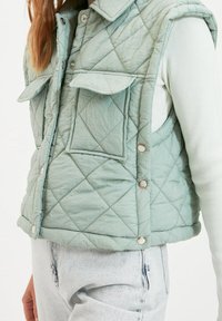 Kamizelka pikowana w jasnym miętowym kolorze, z zapięciem na napy, dwoma przednimi kieszeniami i kołnierzykiem. Teksturowany materiał z przeszytym wzorem diamentowym.