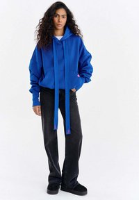 Hanorac albastru supradimensionat cu șnururi lungi, buzunar frontal și manșete ribate, asortat cu blugi negri largi și încălțăminte neagră.