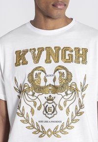 Camiseta blanca con un diseño gráfico dorado que presenta dos pájaros estilizados y elementos decorativos. El texto dice "KVNGH" y "RESUCITA COMO UN FÉNIX."