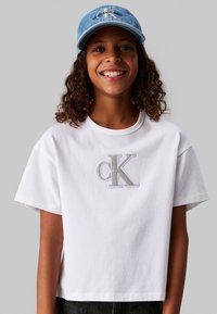 T-shirt bianco in cotone con maniche corte, con logo "ck" ricamato in argento sul davanti. Abbinato a un cappellino di denim azzurro chiaro.