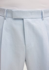 Pantalon bleu clair sur mesure avec passants de ceinture et fermeture avant cachée, porté avec un haut blanc rentré.