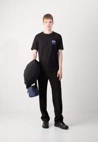 T-shirt en coton noir avec un petit logo violet sur la poche, associé à un pantalon noir et des chaussures noires brillantes ; tenant une veste noire et un sac bleu.