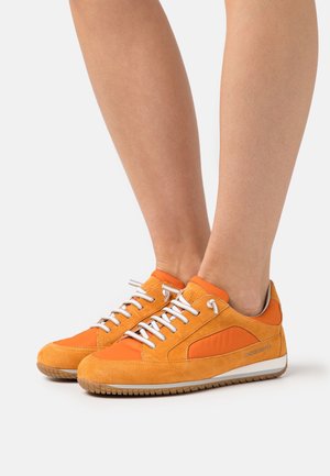 Personne portant des baskets basses en daim orange et tissu avec des lacets blancs et des semelles en caoutchouc, debout sur une surface blanche.