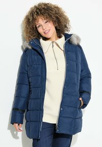 Mörkblå vadderad jacka med huva av päls, dragkedjeslutning, sidofickor och kviltat mönster. Bärs över en krämfärgad tröja.