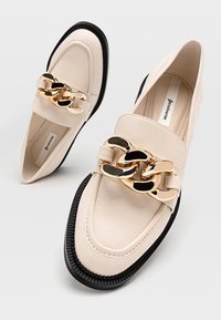 Béžové kožené mokasíny s velkým zlatým a černým kovovým řetězem, s texturovanou černou gumovou podrážkou a zaoblenou špičkou.