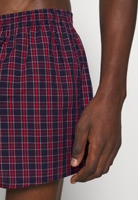Shorts à carreaux bleu marine avec des accents rouges et blancs, dotés d'une taille élastique et d'un matériau en coton léger. Main visible sur le côté.
