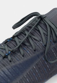 Primo piano di un sneaker lavorato a maglia grigio scuro e blu con lacci testurizzati legati a fiocco su uno sfondo bianco semplice.