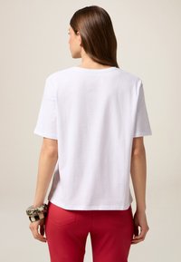 Hvid kortærmet t-shirt med en afslappet pasform, der har en rund halsudskæring. Glat tekstur; båret sammen med røde bukser og dekorative armbånd.