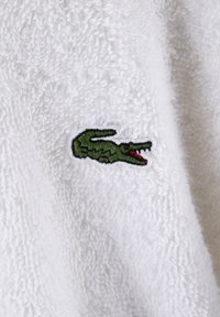 Tissu en éponge blanc avec un logo de crocodile vert brodé et des accents rouges. Texture douce et apparence moelleuse.