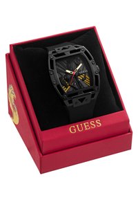 Montre rectangulaire noire avec bracelet en caoutchouc noir, affichant un cadran noir texturé avec des accents dorés et rouges, présentée dans une boîte rouge avec un logo doré.