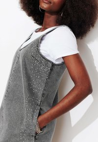 Robe salopette en denim gris à design sans manches, dotée d'une poche et d'études argentées décoratives le long d'un côté. Portée sur une chemise blanche.