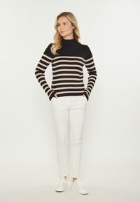 Pulover cu gât înalt, cu dungi negre și crem, cu textură canelată, purtat împreună cu pantaloni slim albi și adidași albi. Modelul stă cu mâinile în buzunare.