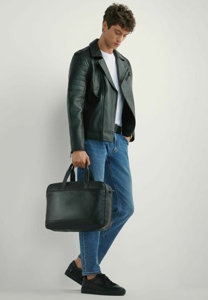 Młody mężczyzna w czarnej skórzanej kurtce, białej koszulce, niebieskich dżinsach i czarnych trampkach, trzymający czarną skórzaną teczkę.