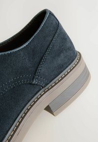 Chaussure en daim bleu foncé avec un petit talon, présentant des détails de couture et une semelle beige contrastante. La tige a une texture lisse.