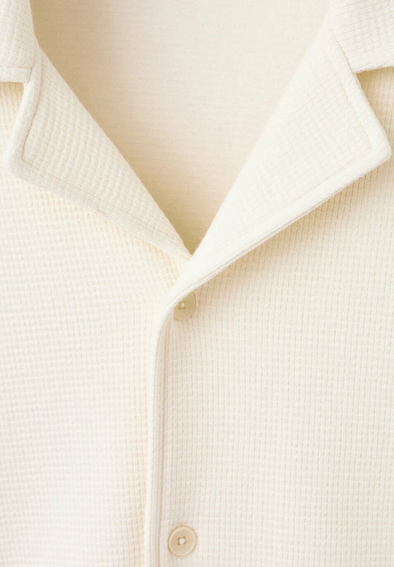 Primer plano de camisa abotonada texturizada de color crema con cuello puntiagudo y dos botones visibles.