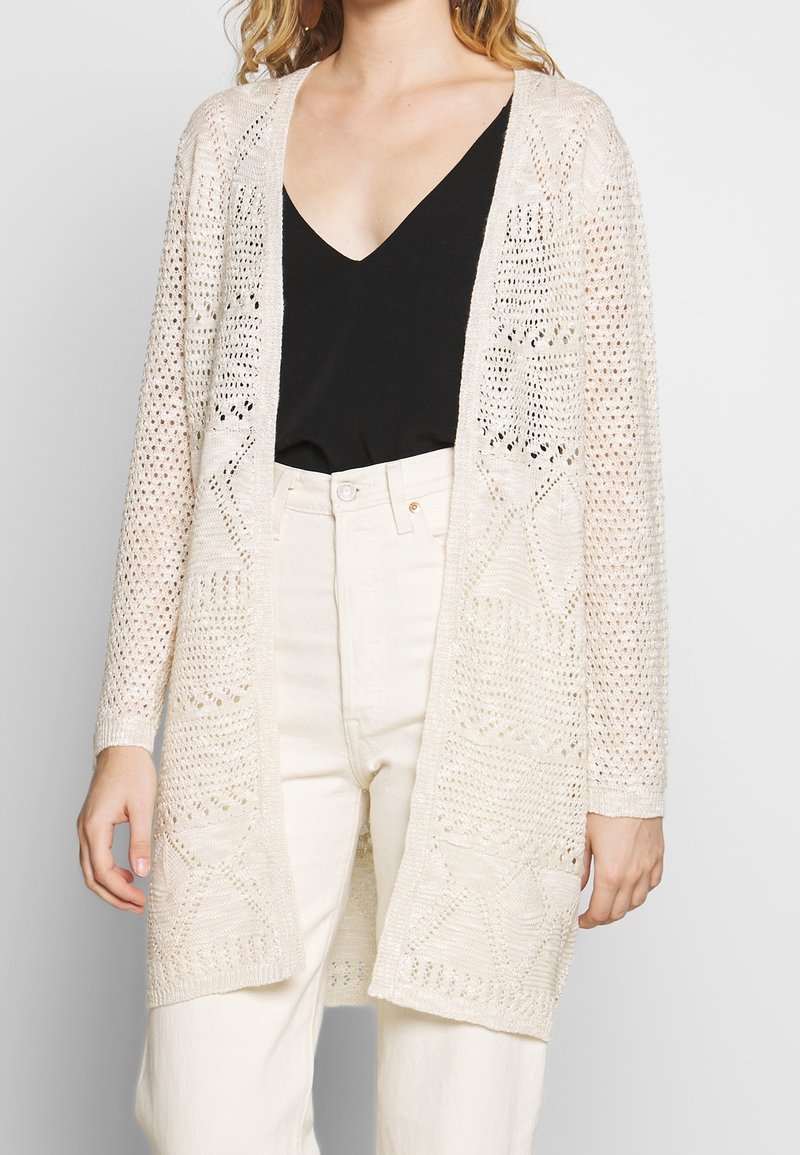 Strickjacke in Hellbeige mit offenem Vorderteil, mit gemustertem Strukturdesign und langen Ärmeln. Trägt man über einem schwarzen Oberteil und hellen Hosen.