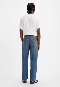 Personne aux cheveux tressés portant un t-shirt blanc, un jean droit bleu et des baskets noires, debout de dos sur un fond blanc.