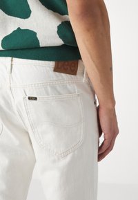 White denim jeans with stitched back pockets and a brown leather label. Sweater features green and white abstract pattern.