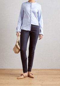 Ljusblå skjorta med volangärmar, marinblå tajta byxor och beige slip-on skor. Individuen har en beige väska. Bakgrunden är en träffloor.