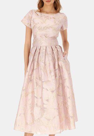 Kobieta ubrana w sukienkę z różowo-złotym kwiatowym żakardem, z krótkimi rękawami, dopasowaną talią i plisowaną, sięgającą do kostek spódnicą, stojąca z jedną ręką w kieszeni.