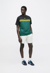 T-shirt verde e preta com riscas amarelas, combinada com calções claros e ténis azuis. Design simples e casual com mangas curtas.