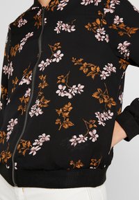 Svart bomberjacka med rosa och bruna blommönster, ribbade muddar och midja, öppen dragkedja, mjuk tygtextur, åtsittande design.