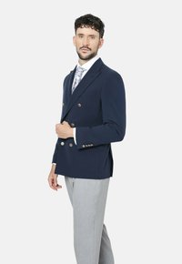 Blazer azul marino de doble botonadura con botones dorados, con un tejido texturizado y un corte entallado, combinado con pantalones gris claro y una corbata estampada.