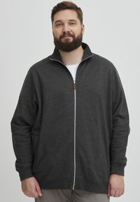 Felpa grigia con zip e colletto alto, caratterizzata da una sottile texture, zip bianca e un'etichetta con logo, indossata sopra una maglietta bianca.