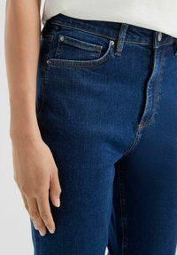 Temno jeans tkanine z tesno prilegajočim dizajnom, ki vključuje vidne šive, sprednje žepe in gladko teksturo. Delno vidna roka ob strani.