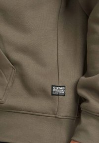 Sudadera verde oliva con textura acanalada, bolsillo tipo canguro y etiqueta del logo de G-Star Originals en la parte inferior izquierda.