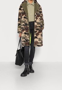 Płaszcz moro z futerka syntetycznego w brązowych i zielonych tonach, zestawiony z jasną zieloną bluzką z zamkiem przy kołnierzu, czarnymi jeansami i czarnymi butami typu chelsea, trzymający czarną torbę.